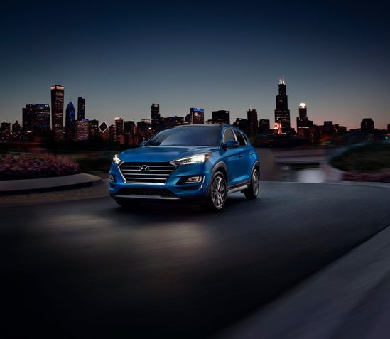 blue Tucson driving at night with headlights on with city in background