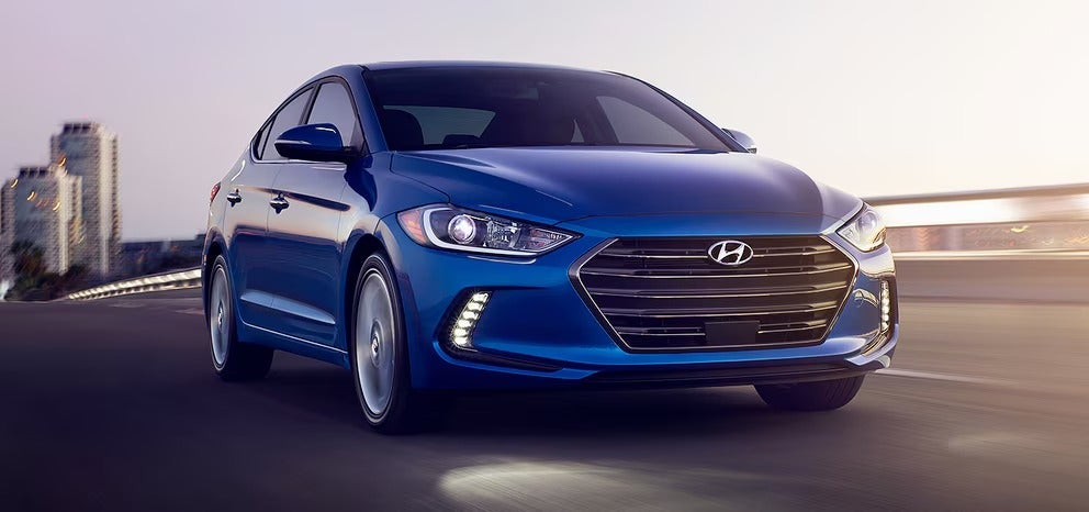 blue Elantra driving on a highway at dusk with a city in the background