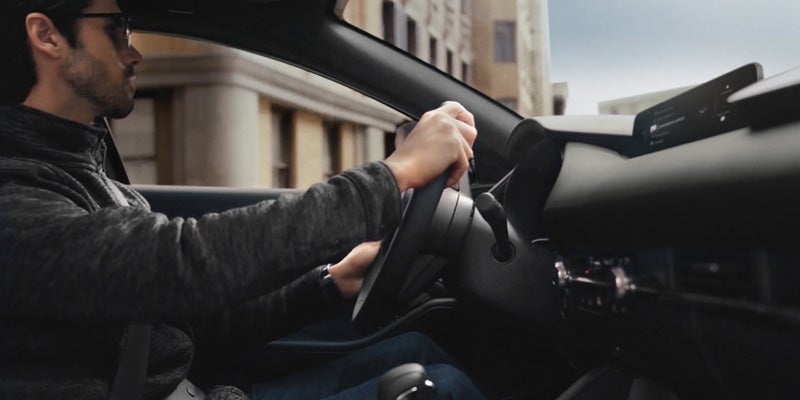Side profile view of a man with sunglasses and a dark fleece pullover driving a car, with his hands positioned on the steering wheel inside a modern vehicle cabin.