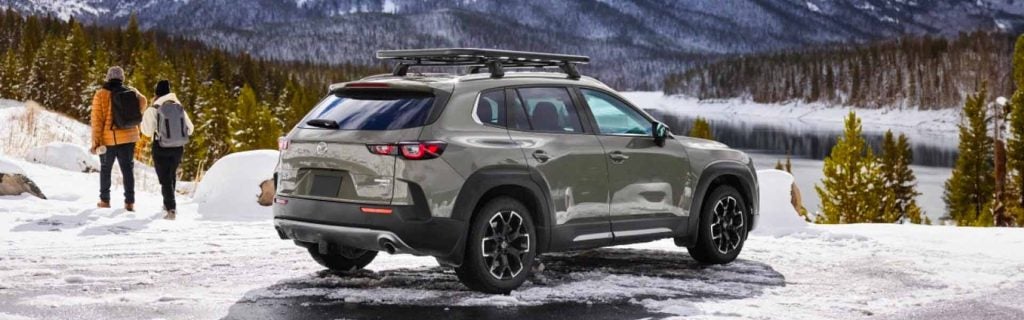 A green Mazda CX-50 SUV with a roof rack parked on snow near a lake, with two backpackers walking toward a snowy mountain landscape.