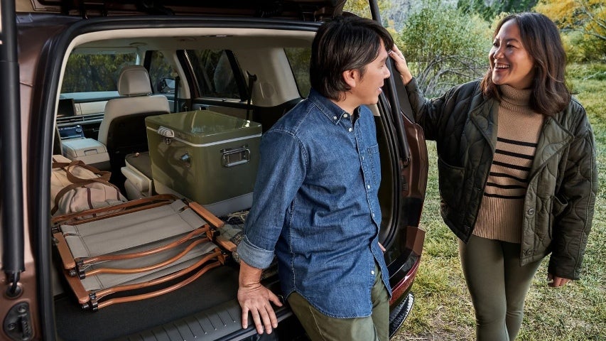 Man and woman smiling beside open SUV trunk packed with camping gear including a cooler, folding chairs, and backpacks.