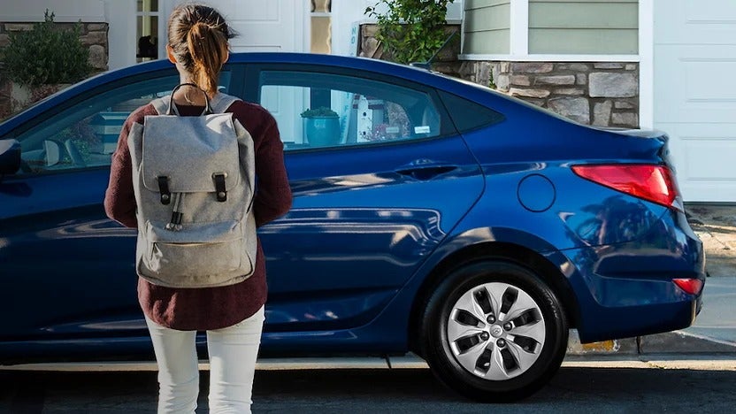 Young woman with a gray backpack standing in front of a parked blue sedan outside a suburban home.