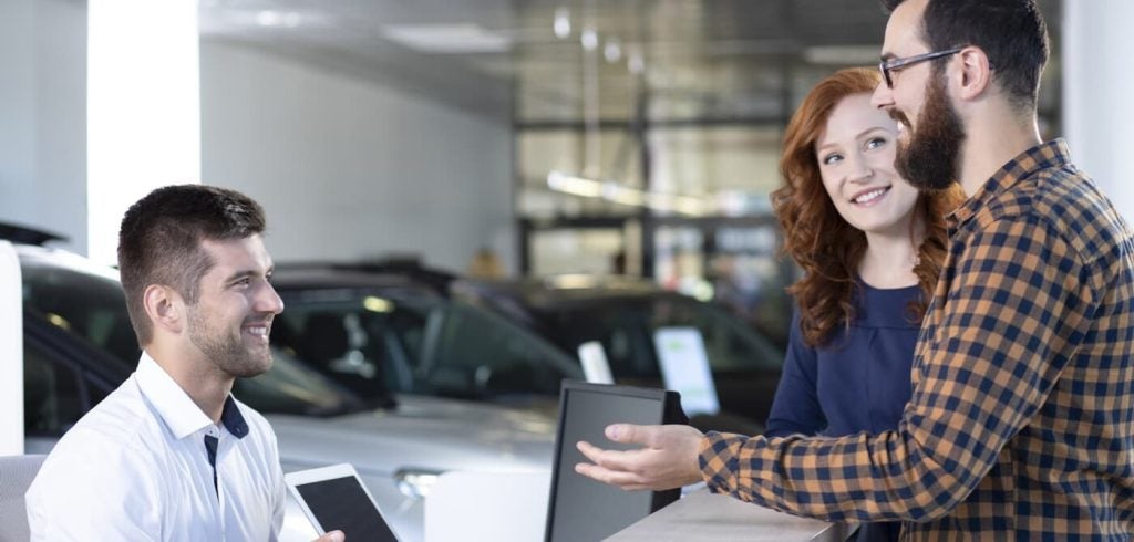 Three people smiling while looking at a car in a dealership.