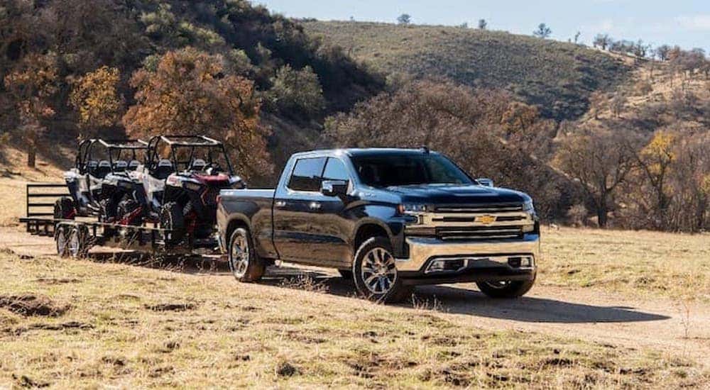 A black 2020 Chevy Silverado 1500 towing a trailer with ATVs on a dirt path.