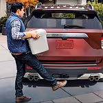 A person in casual attire uses a foot motion to open a red SUV's trunk, holding a white cooler. The scene is set on a city street with autumn foliage.