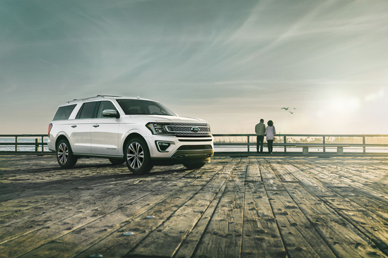 A white SUV is parked on a wooden pier under a serene, cloudy sky. Two people stand in the distance by the railing, overlooking the water, evoking tranquility.