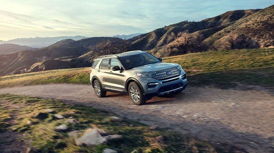 A silver SUV drives on a dirt road through a scenic, hilly landscape under a blue sky, conveying a sense of adventure and freedom.