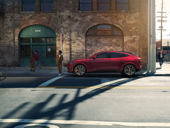 A sleek red SUV is parked on an urban street, casting shadows in the sunlight. A brick building in the background, people walking, and a sense of calm.