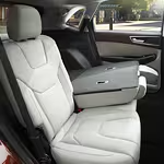 Rear interior of a car featuring plush gray seats, one folded down as an armrest with cup holders. Bright exterior visible through windows. Luxurious and modern.