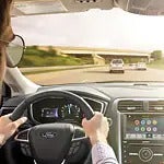 A person with sunglasses drives a car on a sunny highway. The dashboard displays navigation apps. Two cars are ahead, with a bridge in the distance.