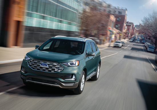 A sleek, green SUV drives down an urban street lined with modern and historic buildings, under a clear blue sky. The scene conveys motion and urban energy.
