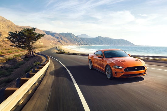 Orange 2018 Ford-Mustang-GT-Coupe driving on a coastal highway, with ocean on the right and rugged hills on the left under a blue sky, conveying freedom and adventure.