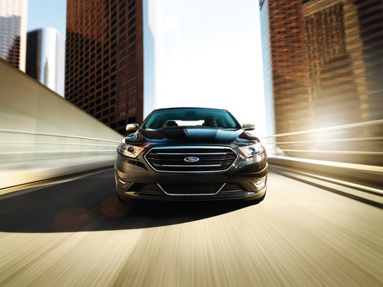 A black car drives quickly on a city bridge, creating a sense of speed. Tall buildings blur in the background under a bright sky, highlighting urban energy.
