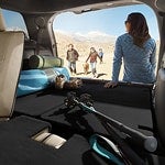 A woman in blue stands by an open car trunk filled with hiking gear, overlooking a group walking in a sandy, mountainous area under a clear sky.
