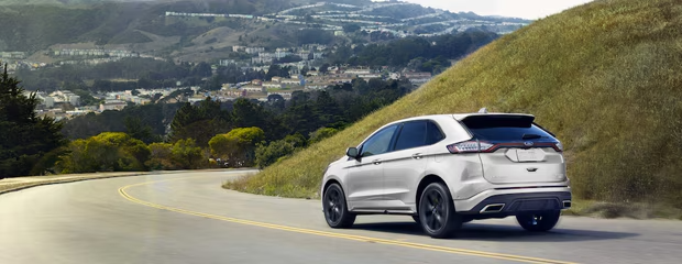 A white SUV drives along a winding road through lush hills on a sunny day. In the background, a scenic view of a town and distant mountains unfolds.