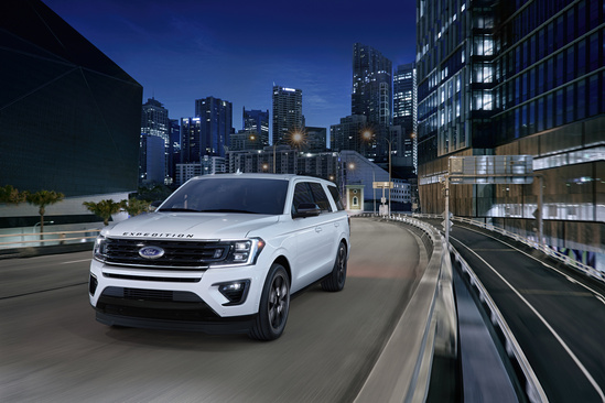 A white SUV drives on an elevated road through a modern city at night. Skyscrapers with glowing lights line the background, conveying a sleek, urban vibe.