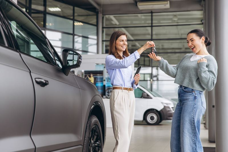 A woman handing car keys to another woman in a car dealership.