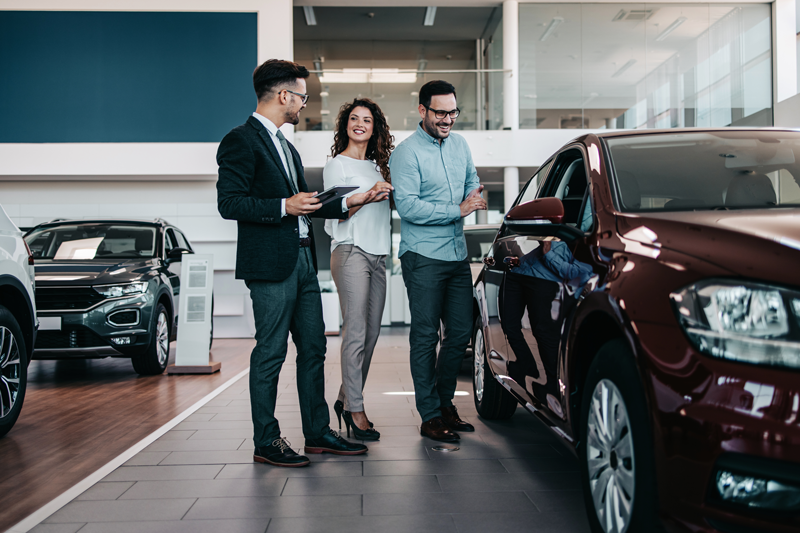 Salesperson showing a couple a maroon sedan in a dealership showroom, discussing features beside the car.