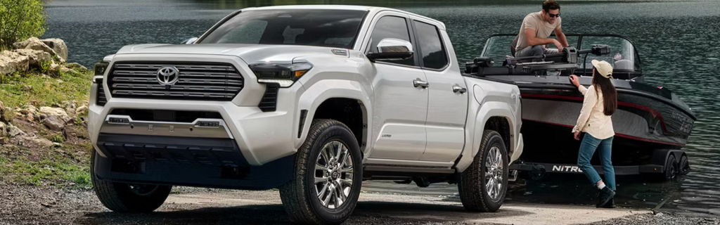 A white Toyota Tacoma truck parked by a lake, with a person sitting in the truck and another person standing next to a boat.