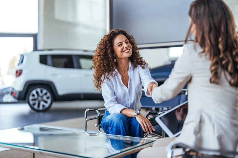 Smiling customer shaking hands with a sales representative in a modern car dealership showroom, seated beside a glass desk with a vehicle visible in the background.