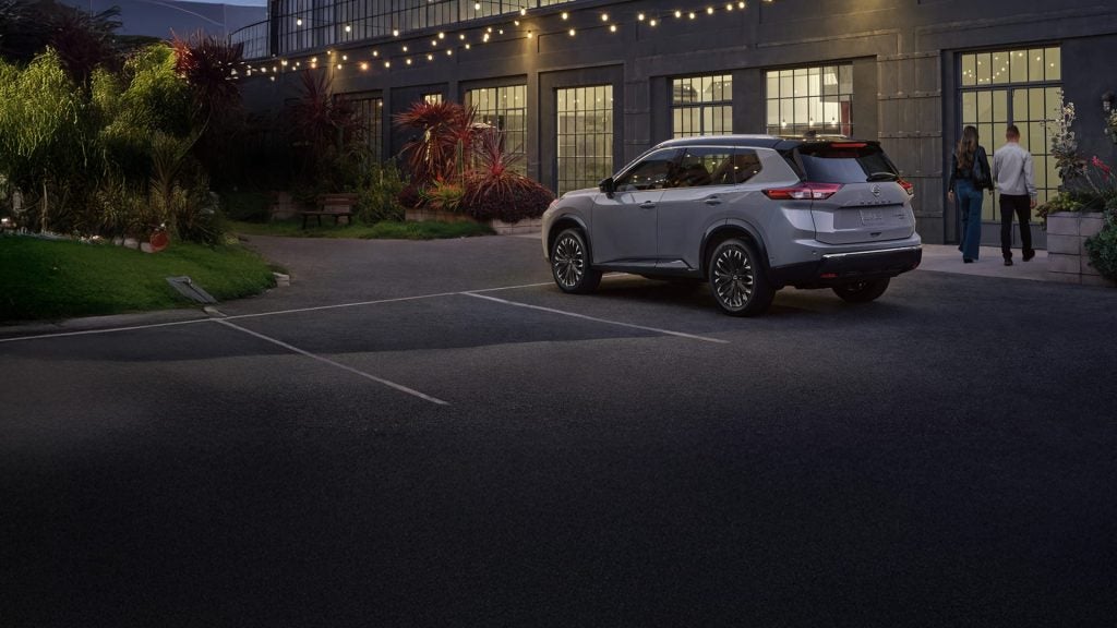A Silver Nissan SUV parked outside a modern building at night with string lights.