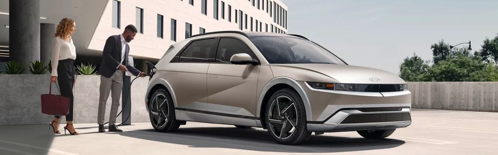 A man in a suit is charging a Hyundai IONIQ 5 electric vehicle while a woman with a bag looks on, set in an urban environment with buildings in the background.