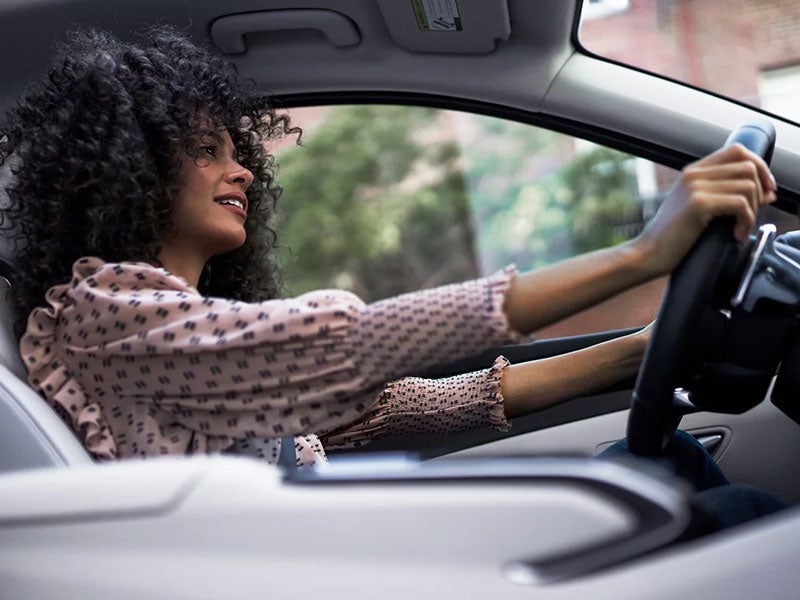 Woman with curly hair smiling while driving a car and holding the steering wheel.