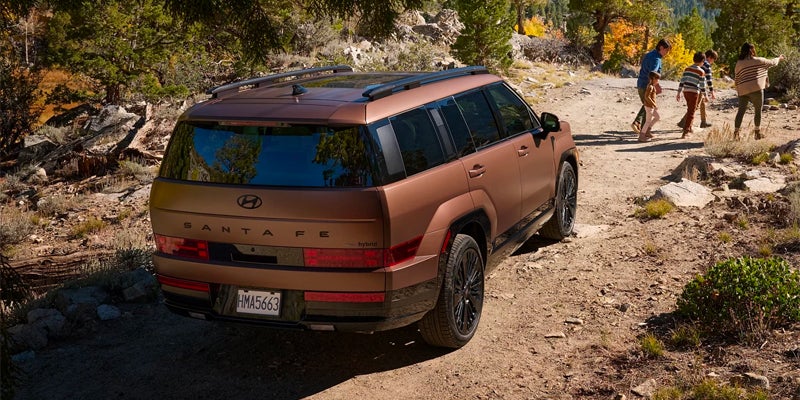 Hyundai Santa Fe Hybrid SUV driving on a rugged forest trail, with a family walking ahead among trees and rocky terrain during an outdoor adventure.