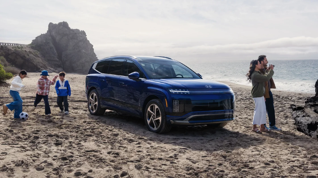 A blue IONIQ9 SUV parked on a beach with a rocky coastline. A family of five is nearby: two adults are taking a selfie while three children, one of whom is holding a soccer ball, are playing in the sand.