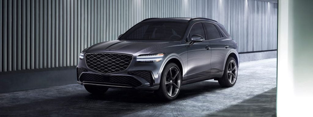 Sleek gray 2026 Genesis GV70 parked in a modern industrial setting, featuring a bold diamond-pattern grille, slim LED headlights, large alloy wheels, and a refined, aerodynamic design.