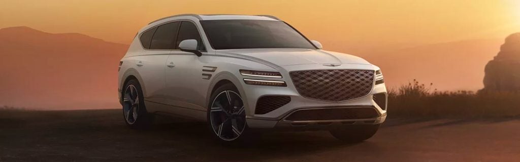 White 2026 Genesis GV80 luxury SUV driving on an open road at sunset with a bold crest grille and LED headlights.