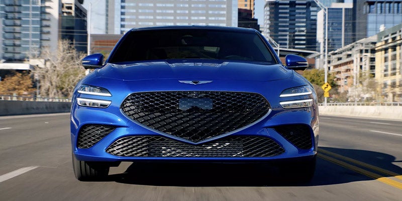 The front view of a blue 2026 Genesis G70 navigating a city street, highlighting its modern design and urban environment.