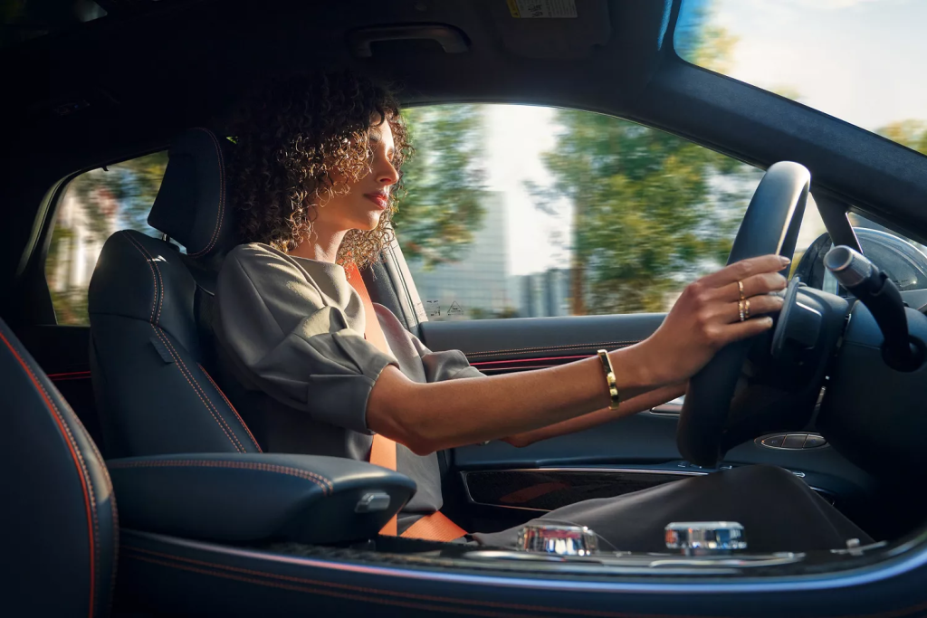 A woman with curly hair driving a Genesis car, focused on the road with her hands on the steering wheel.