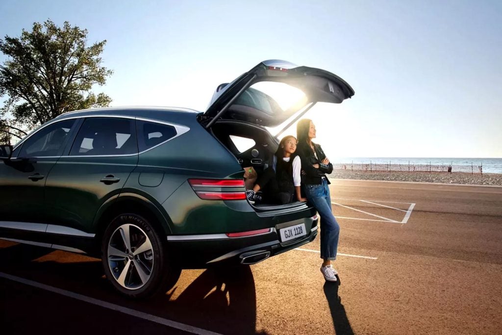 Genesis luxury SUV parked by the beach with the rear liftgate open as two people enjoy the ocean view at sunset.