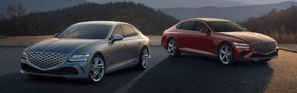 Two 2026 Genesis G80 cars positioned in front of towering mountains, showcasing a scenic view.