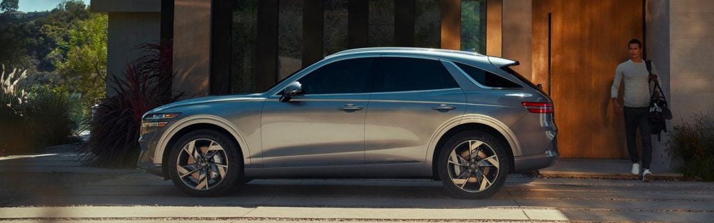 Side profile of a silver 2026 Genesis GV70 luxury SUV parked outside a modern home with sleek alloy wheels and LED taillights.