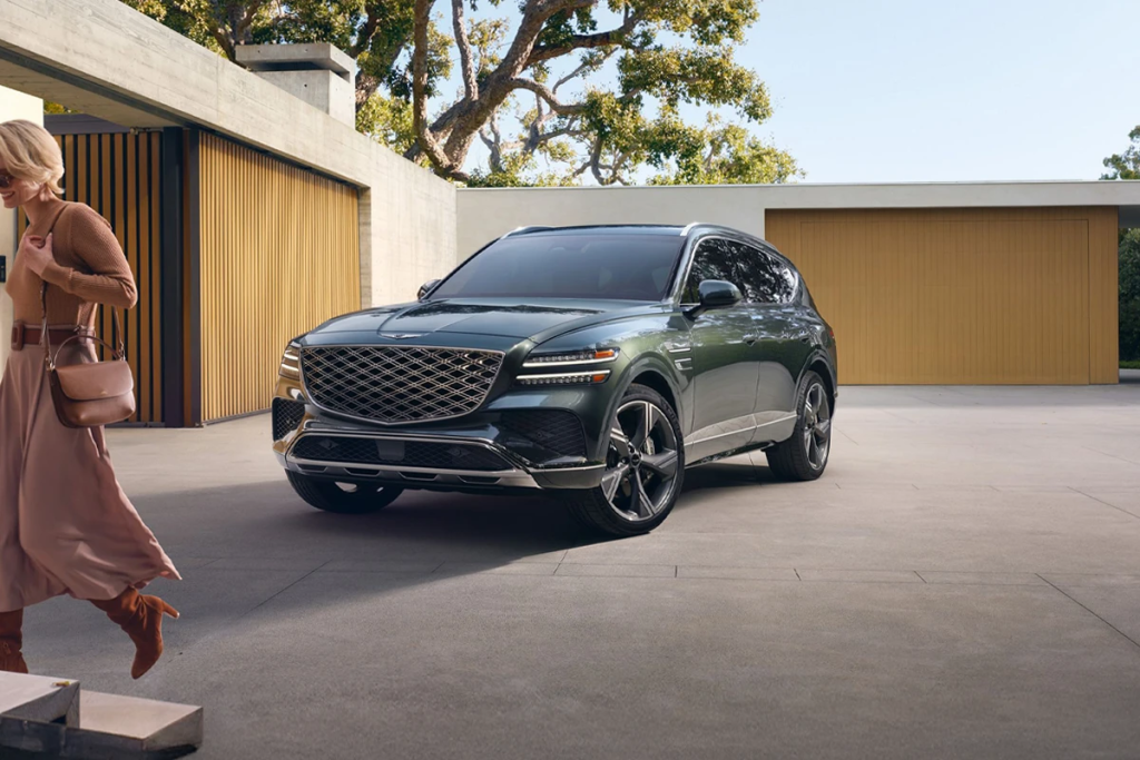 Luxury green Genesis SUV parked in a modern driveway, featuring a bold grille, sleek LED headlights, and a stylish contemporary home backdrop with a woman walking nearby.