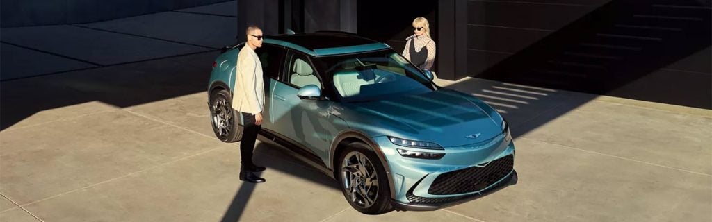 A stylish 2026 Genesis GV60 SUV parked in an outdoor setting with two people standing beside it, one on each side.