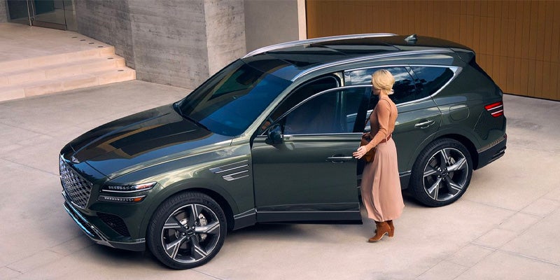 A woman in a long dress and boots stands next to the open driver's door of a dark green Genesis GV80 SUV parked in a modern driveway.