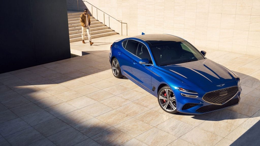 A blue Genesis luxury car parked on a tiled surface with a person walking in the background.
