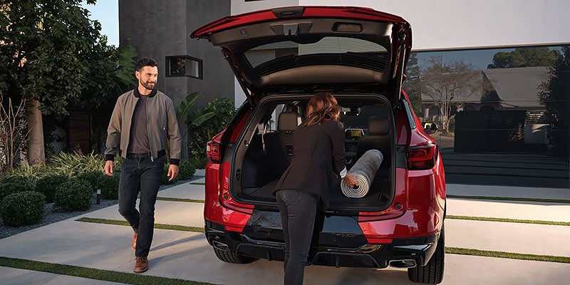 A man stands outside a red Chevrolet Blazer SUV while a woman crouches inside the open trunk, handling a rolled item. The setting is modern, with greenery and a contemporary building in the background.