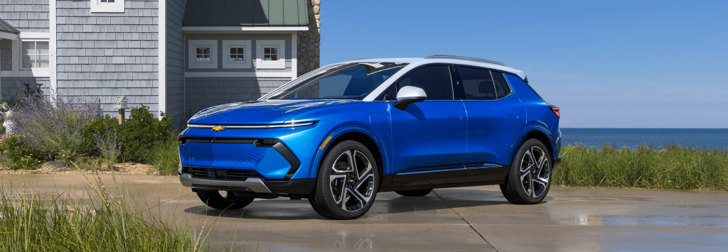 Bright blue Chevrolet electric SUV parked on a seaside driveway beside a gray coastal-style home, with grassy dunes, ocean views, and a clear blue sky in the background.