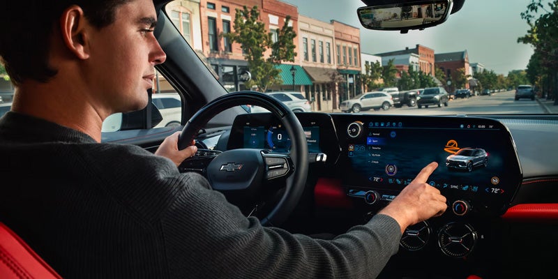 A person sitting in the driver's seat of a car, interacting with a touchscreen dashboard while driving through a city street.