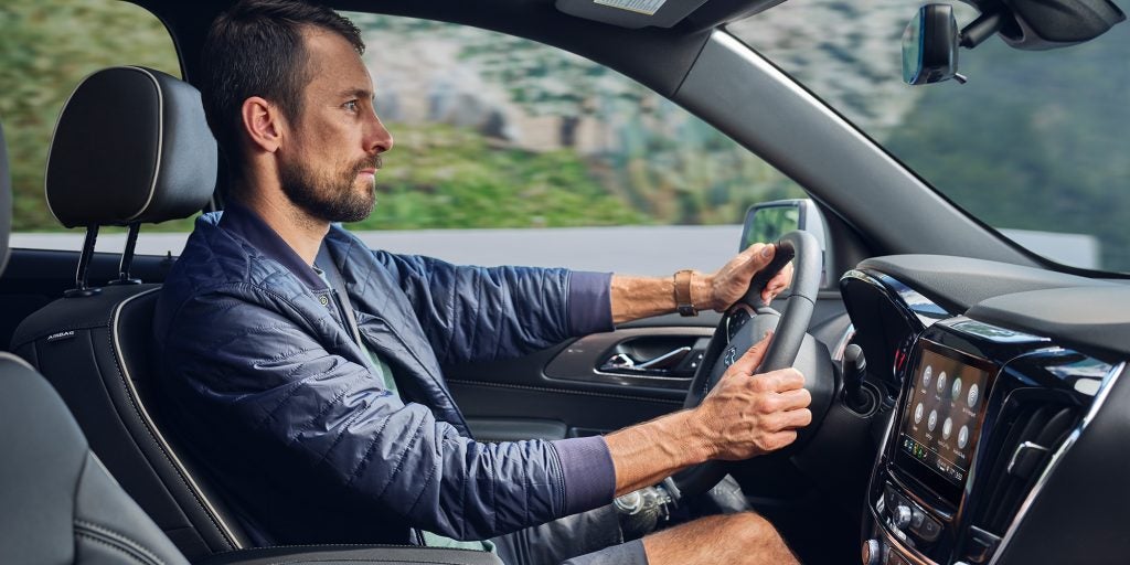 Driver secured with a seatbelt, both hands gripping the steering wheel, eyes forward, with visible side mirror and an accessible infotainment screen
