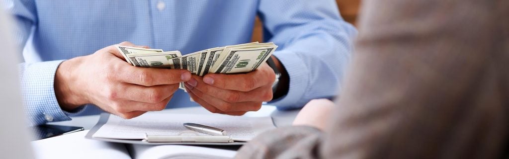 A person holding a stack of cash while seated at a table, with another person's hands partially visible in the foreground.