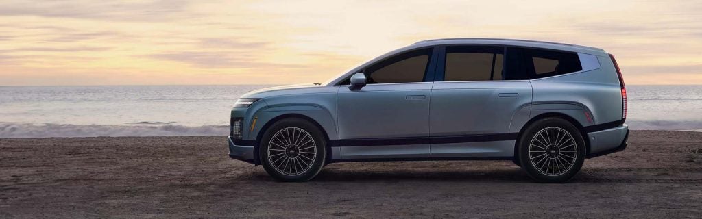 Silver Hyundai IONIQ 9 electric SUV parked by the beach at sunset, shown in side profile with ocean waves in the background.