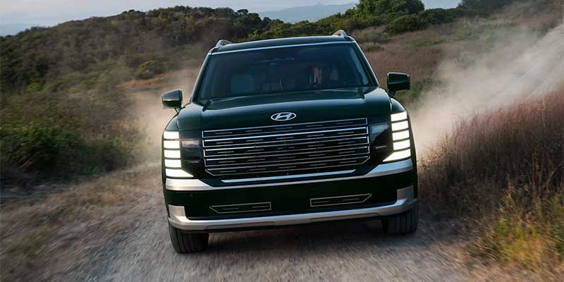 A green Hyundai Palisade driving on a dirt road, showcasing its front design and powerful stance. The vehicle is surrounded by natural terrain, emphasizing its rugged capability.