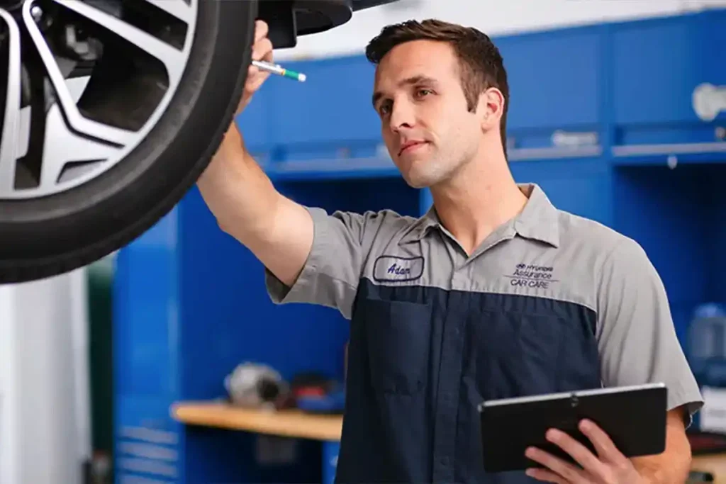 A man in a mechanic's uniform inspecting a car tire while holding a tablet.