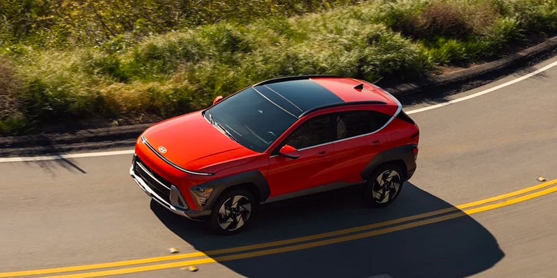 A red Hyundai KONA SUV driving on a winding road surrounded by greenery.
