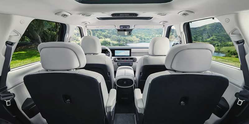 Spacious Hyundai Palisade interior with three rows of light-colored seats, panoramic sunroof, and a modern dashboard featuring a central touchscreen, viewed from the rear looking toward the front, with scenic greenery visible through the windows.